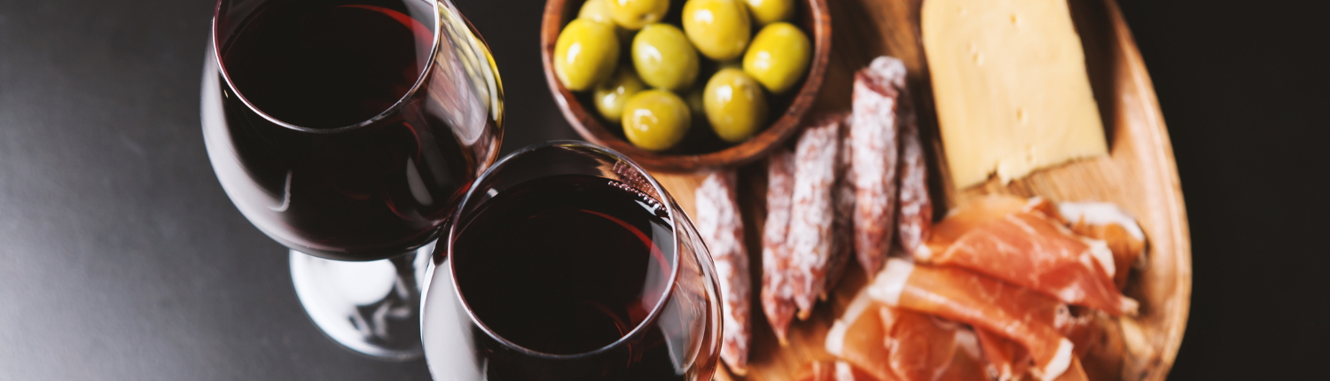 Two glasses of red wine with meat, cheese, and olives in the background.